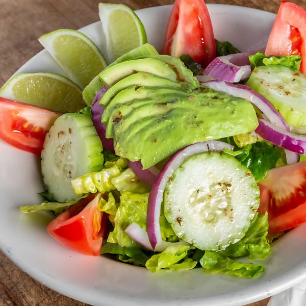 Close-up of a fresh Cuban salad with avocado, tomatoes, cucumbers, red onion, and lime wedges.