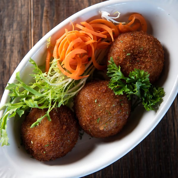 Three golden-brown croquettes with fresh greens and shredded carrots in a white dish on a wooden table.