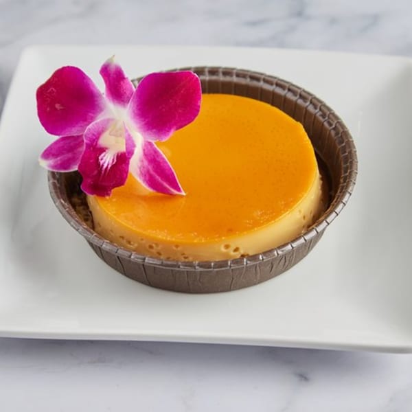 Cuban flan dessert with a vibrant orange top, garnished with a pink orchid, on a white plate.