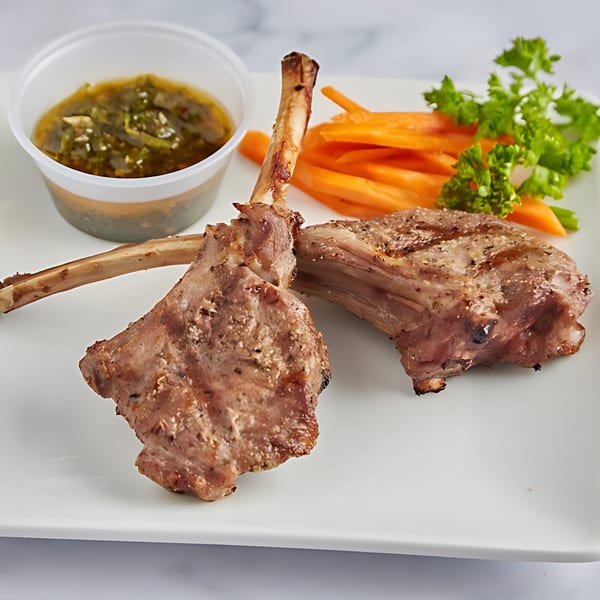 Two grilled lamb chops with carrots, parsley, and a side of green sauce on a white plate.
