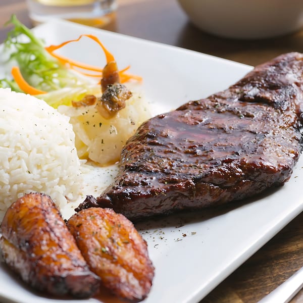 Cuban grilled steak with rice, plantains, and a side salad on a white plate.