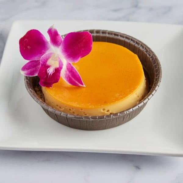 Cuban flan dessert with a vibrant orange top, garnished with a pink orchid, on a white plate.